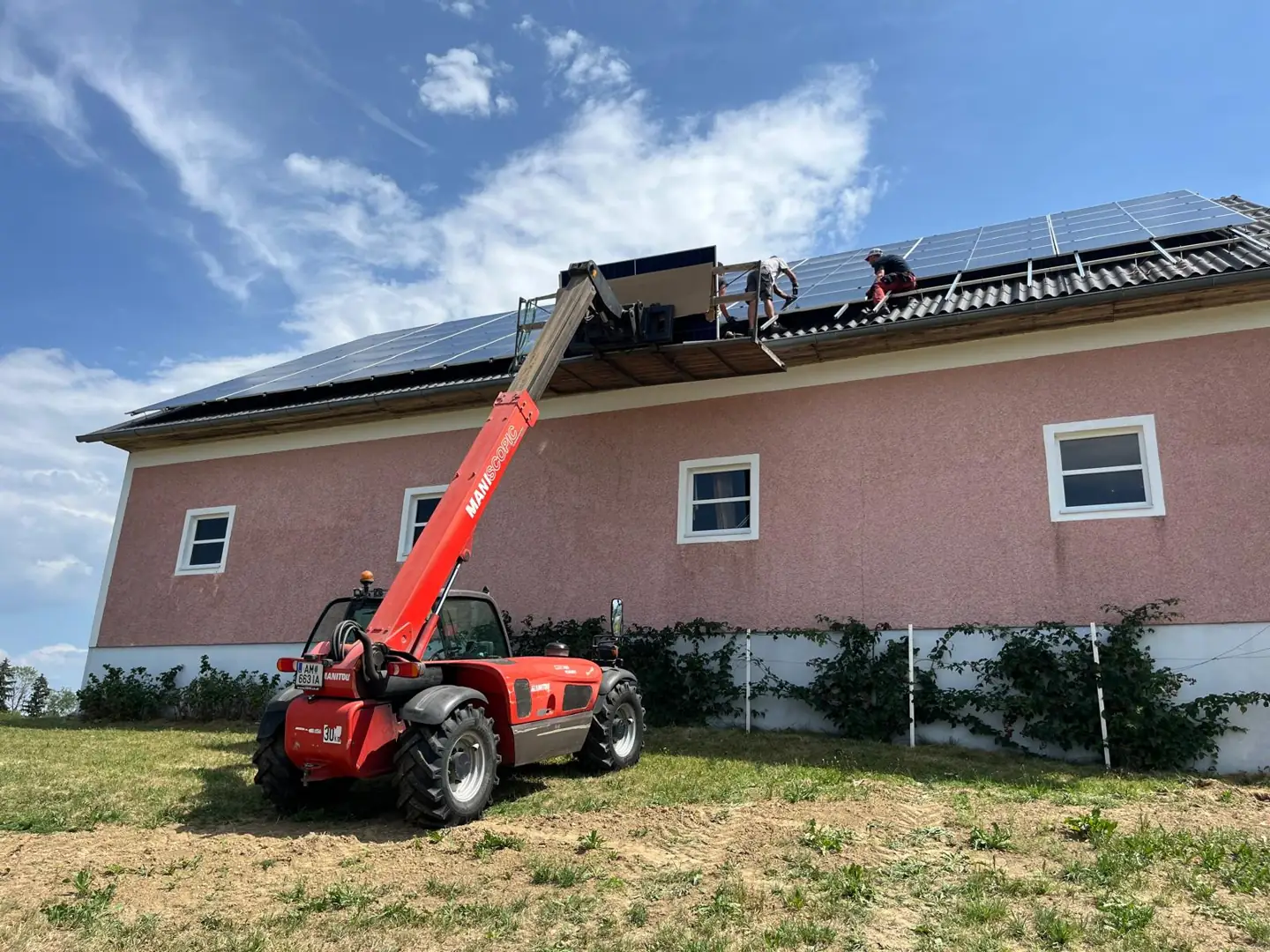 Roter Teleskoplader mit Arbeitsbühne an der Halle von Behamberg Alpakas, zwei Monteure von Elektrotechnik Hochbrandner setzen PV-Module am Dach